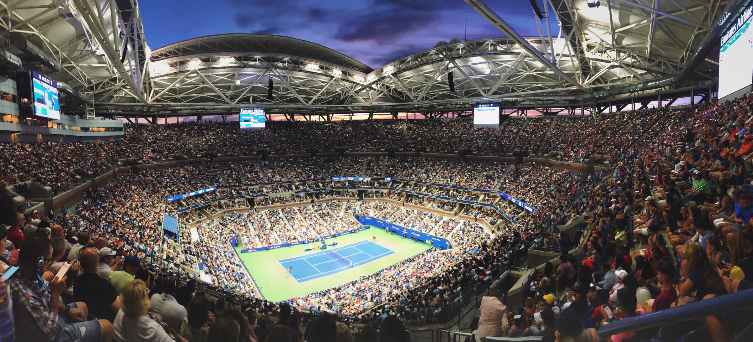 Panorama of a packed Arthur Ashe Stadium at dusk, floodlights on, the roof open above a blue court.